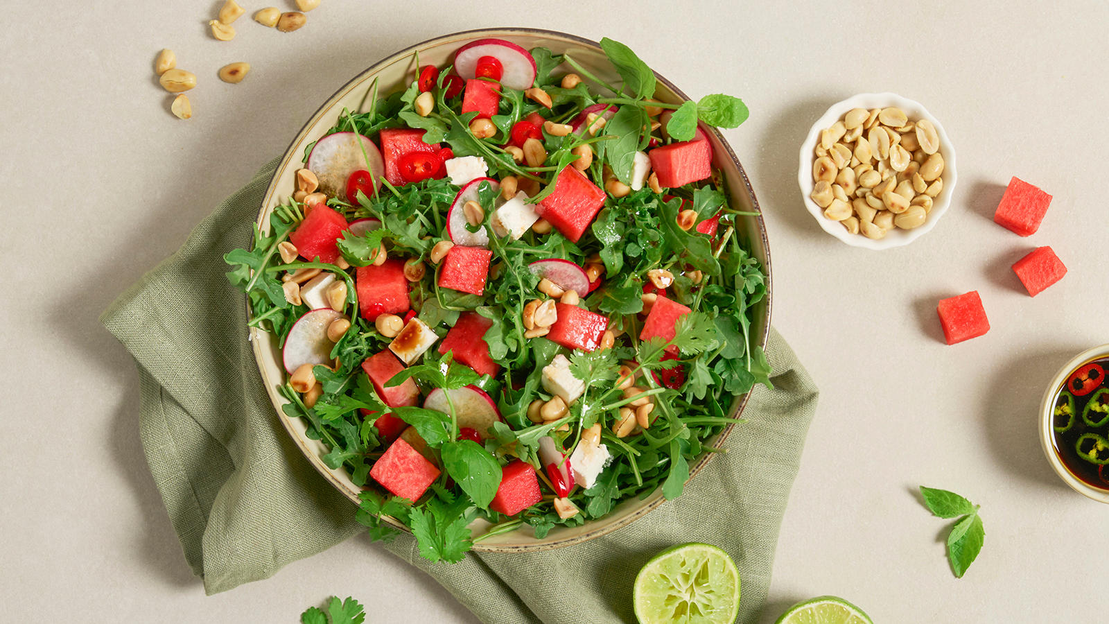 Frisk vannmelonsalat med fetaost og urter servert i lys keramikkskål på grønt kjøkkenhåndkle. Liten skål med peanøtter, og liten skål med dressing på siden.