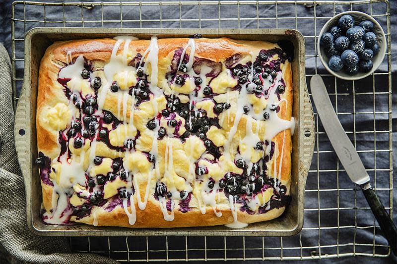 Gylden bollefocaccia i langpanne, med blåbær og vaniljekrem. Melisglasur ringlet over. Mørk bakgrunn, med ferske blåbær på siden.