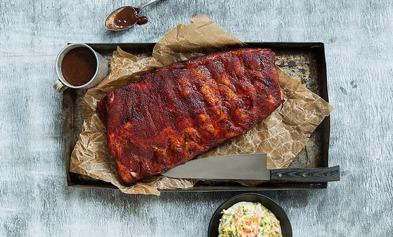 Saftige spareribs servert på metallbrett med bakepapir. Hjemmelaget kålsalat og bbq saus i skåler på siden.