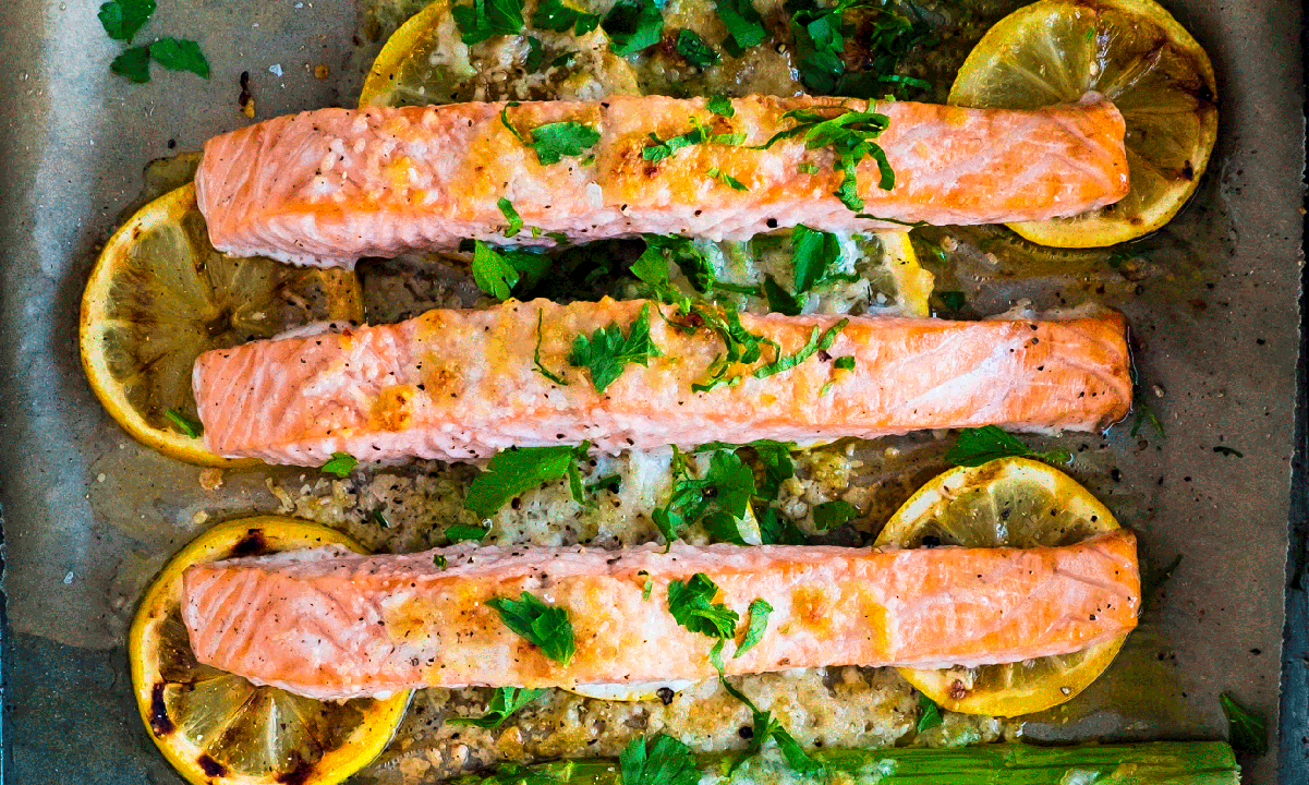 Ovnsbakt laks med parmesan og asparges servert på sitronskiver på bakepapir i ildfast form. Finhakkede friske urter er strødd over fisken.
