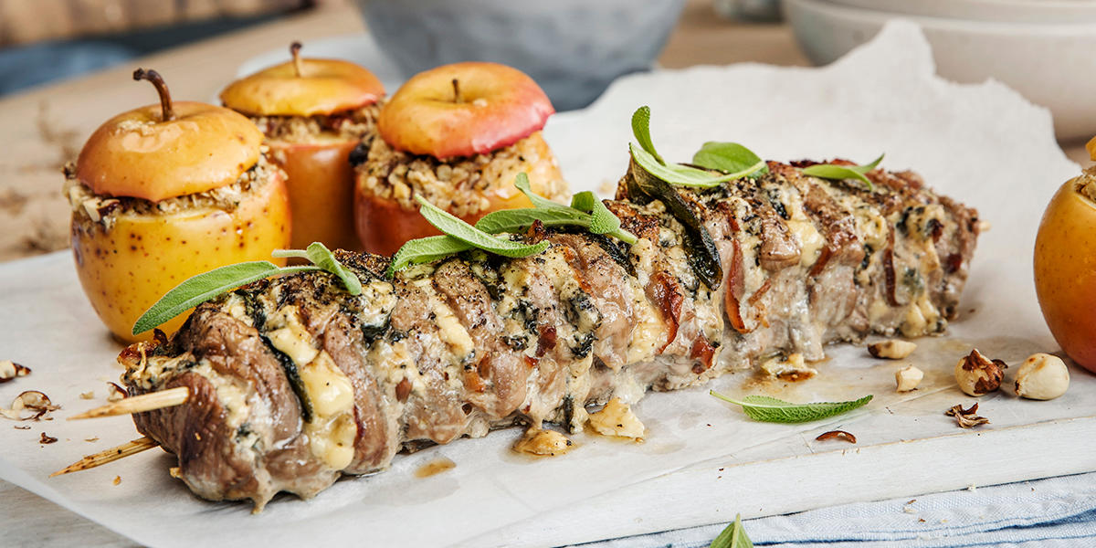 Hasselback svinefilet med blåmuggost, servert på hvit trefjøl, med hvitt bakepapir, overstrødd med fersk salvie. Bakte hele epler med kryddersmør i bakgrunnen.