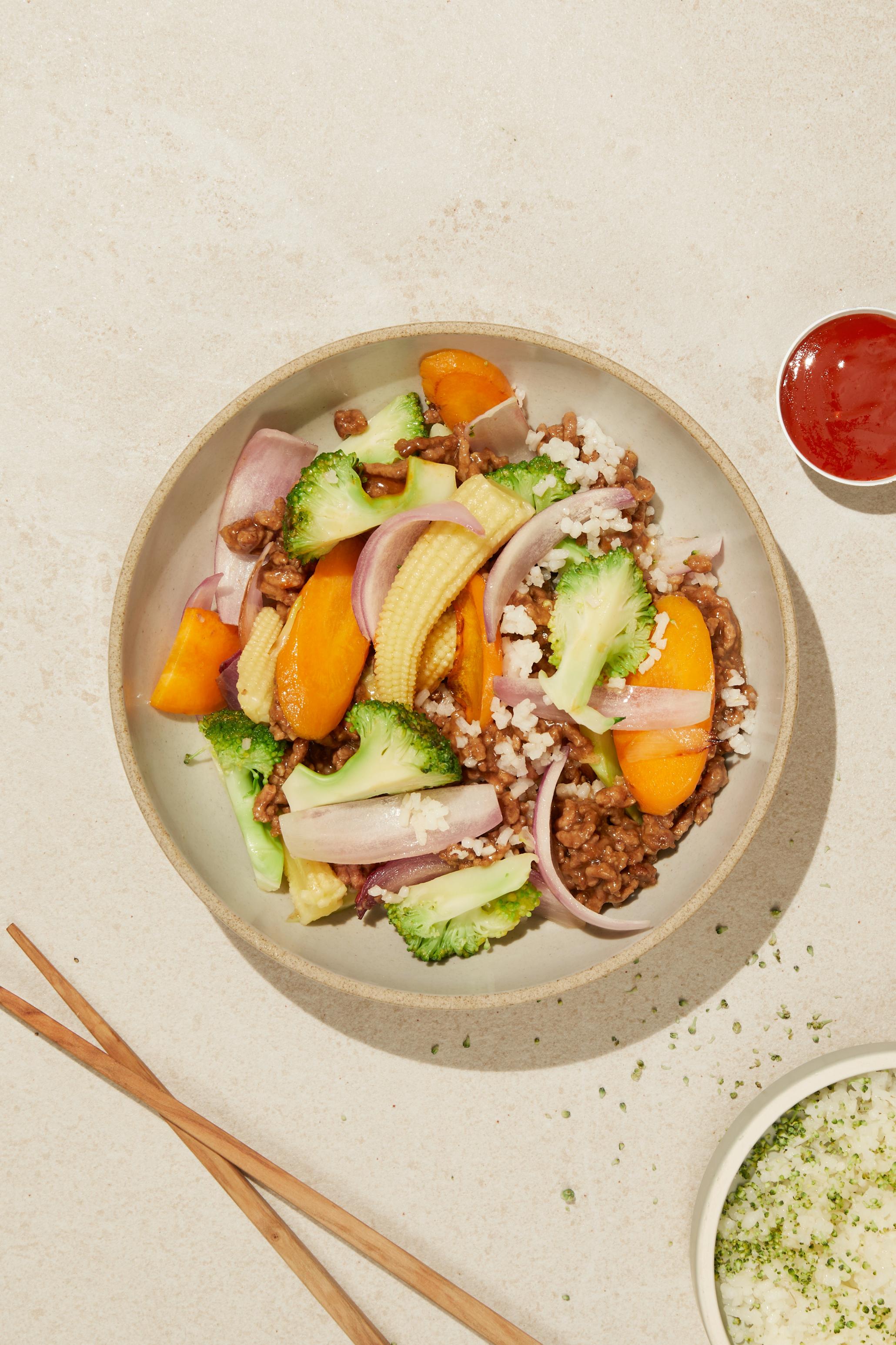 Wok med kjøttdeig og sursøt saus, med brokkoli, gulrot, rødløk og ris. Servert i beige skål, på beige bakgrunn.