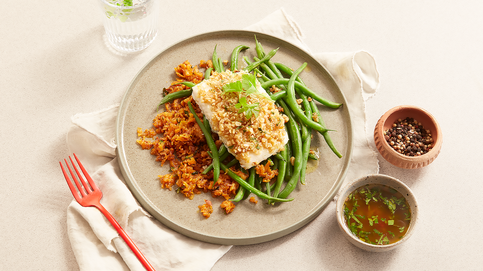 Kveite med nøttelokk og raspet søtpotet servert på lys grå tallerken med kokte grønne bønner. Tallerkenen står på linhåndkle på beige bakgrunn med to skåler krydder og saus ved siden av, og en rød gaffel.