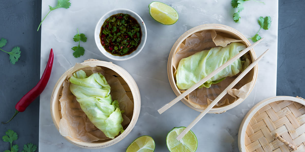 Asiatiske kålruletter med nykål servert i bambuskurver på marmorfjøl. Liten skål med dip ved siden av, sammen med hel rød chili, limebåter og korianderblader.