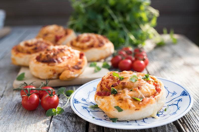 Hjemmelagede pizzasnurrer med tomatsaus og ost. En pizzasnurr på blå og hvit tallerken i forgrunnen, fire pizzasnurrer på trefjøl i bakgrunnen. Ferske urter og tomater rundt.