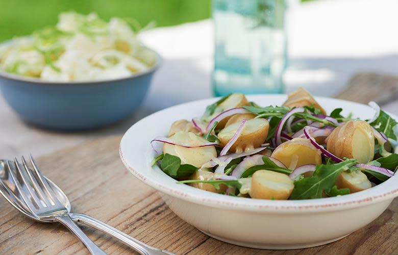 Fransk potetsalat med ruccola og rødløk, og hjemmelaget vinaigrette med honning og sennep. Servert i hvit skål på utendørs sommerbord.
