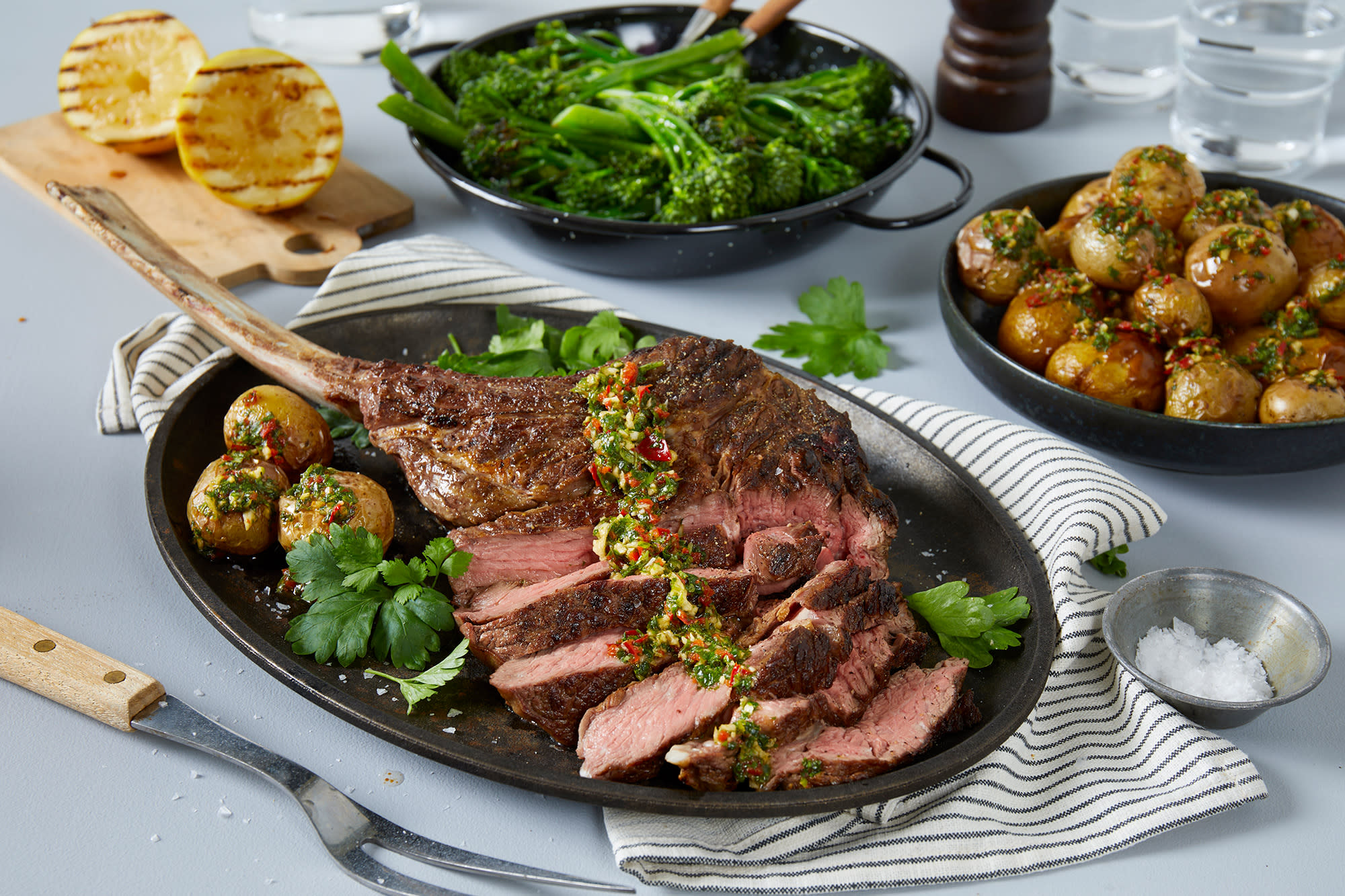 Langtidsgrillet tomahawk med chimichurri poteter servert på mørkt fat med ekstra chimichurri over kjøttet, og grønne urter på siden. Grillet brokkolini, grillet sitron og skål med chimichurri poteter i bakgrunnen.
