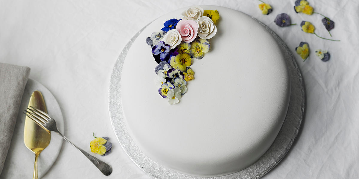 Hvit marsipankake på hvitt fat, pyntet med hjemmelaget kakepynt av kandiserte fioler og marsipanroser. Kakespade og gaffel på siden.