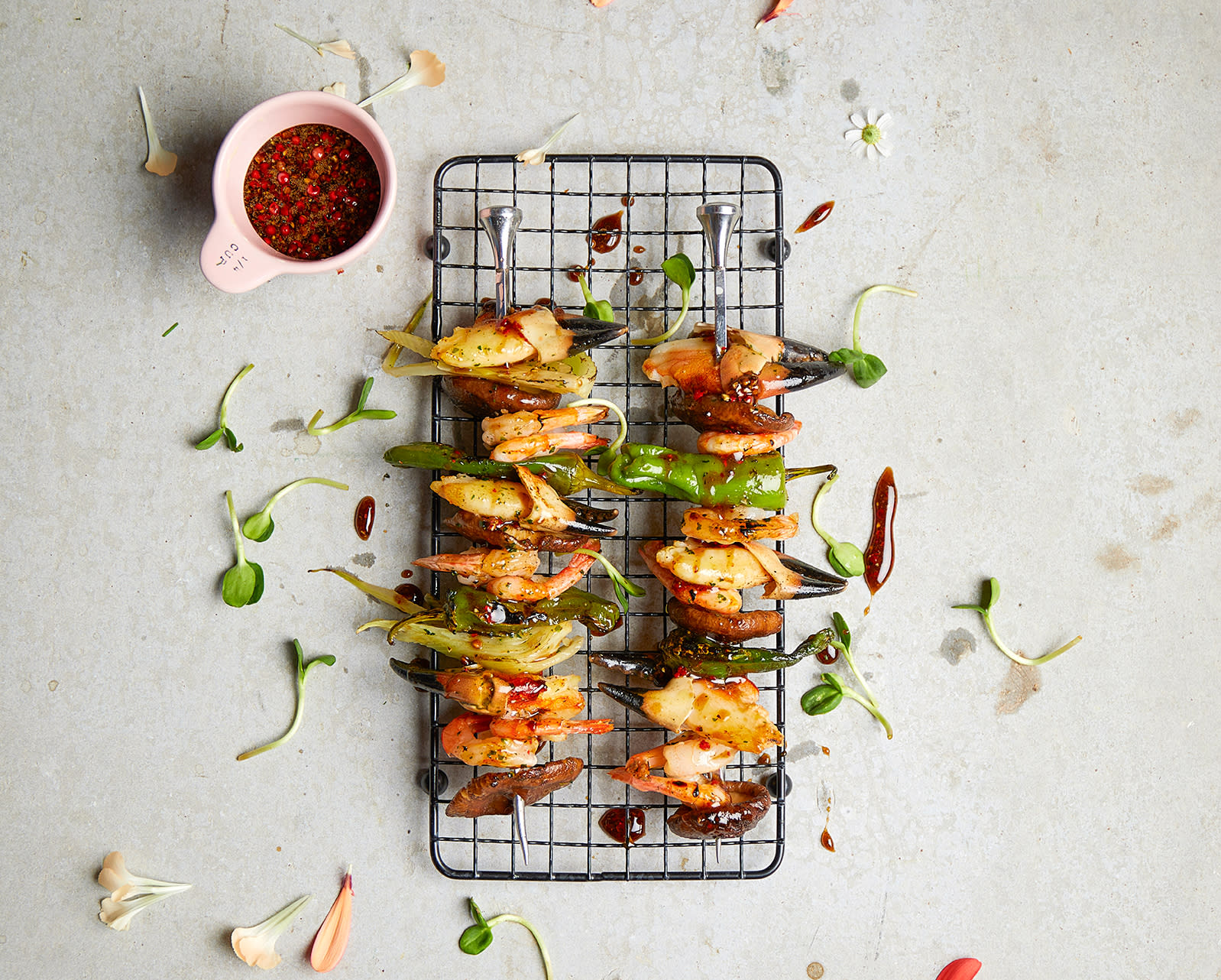 Grillspyd med skalldyr; krabbeklør, reker, villreker, sopp og fennikel, servert på smak rist på lys bakgrunn med friske urter rundt og en liten skål med marinade ved siden av.