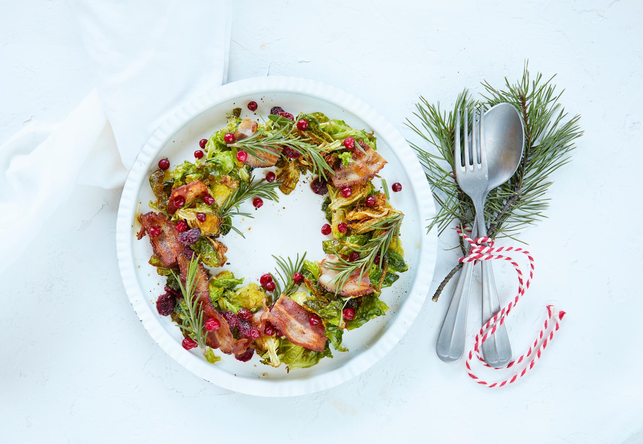 Stekt rosenkål med tranebær og bacon, dandert i sirkel på hvitt fat så de ser ut som en julekrans. Kransen er pyntet med ferske urter, og det er julepyntet bestikk ved siden av.