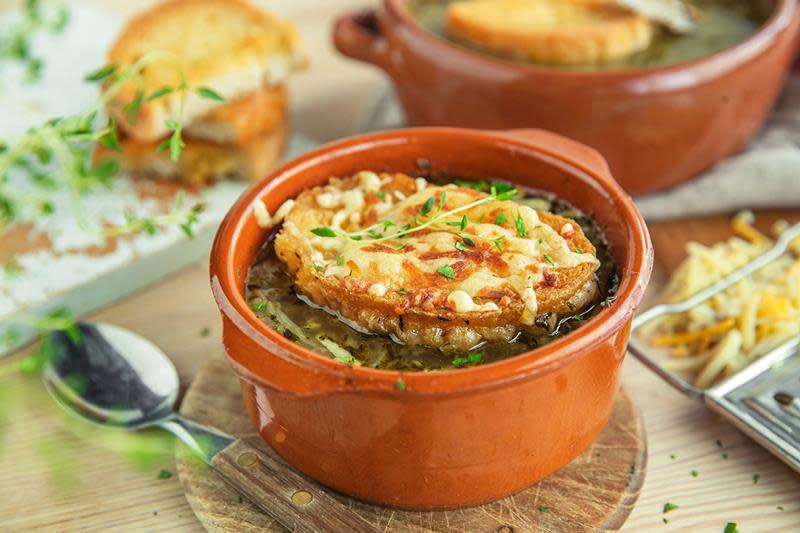 Fransk løksuppe i keramikkskål, på rund trefjøl. Suppen er toppet med ostegratinert brødskive og fersk timian. Ferske urter og ristet brød i bakgrunnen.