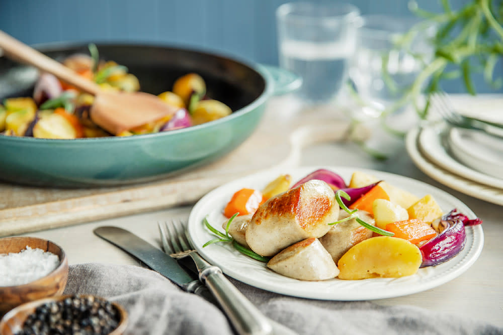 Julepølse med honningglaserte rotgrønnsaker servert på hvit tallerken på dekket middagsbord. Turkis stekepanne med honningglaserte rotgrønnsaker i bakgrunnen.
