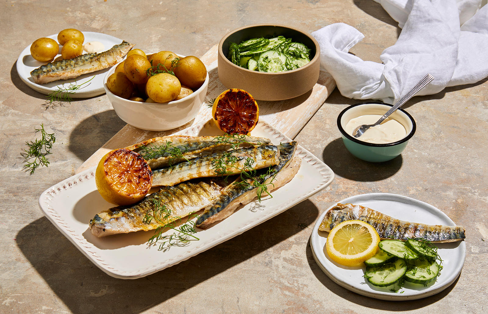 Grillet makrell med agurksalat og nypoteter servert på hvitt firkantet fat, med grillede halve sitroner og frisk dill på toppen. Skåler med nypoteter, agurksalat og rømme ved siden av.