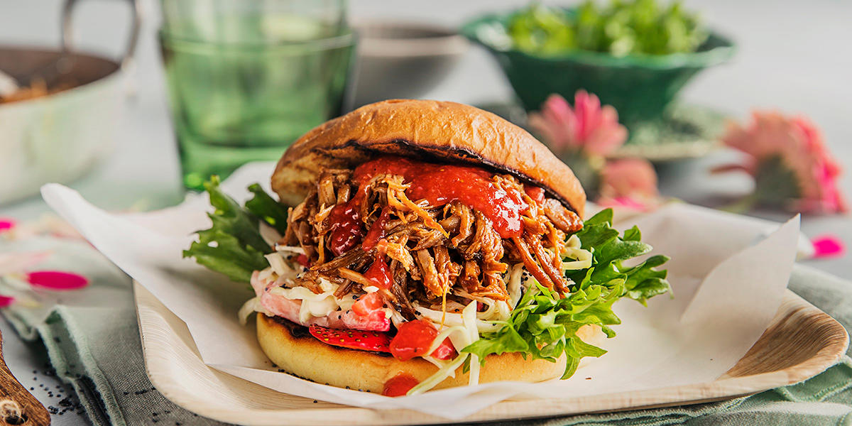 Pulled pork burger med jordbær bbq-saus og fruktig coleslaw servert i grillet burgerbrød med salat, på bambustallerken med bakepapir. Tallerkenen står på dekket middagsbord med en skål salat i bakgrunnen.