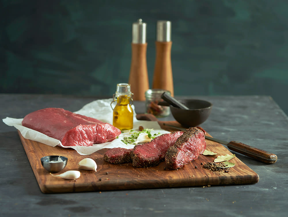 Rå biff i stykke og skiver, med grovkvernet pepper på siden av skivene, servert på slakterpapir på mørk trefjøl med krydder og hvitløk rundt.