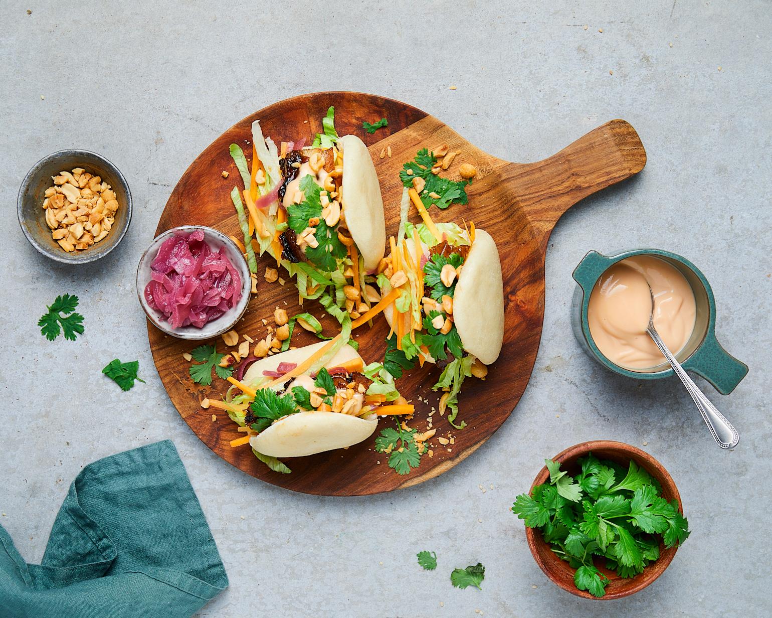 Tre steam buns med glasert ribbe og syltet rødløk servert på rund trefjøl, pyntet med frisk koriander og hakkede peanøtter. Små skåler med chilimayo, frisk koriander og hakkede peanøtter rundt fjøla.