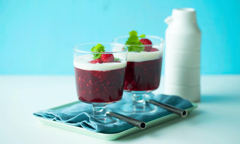 Rødgrøt med bringebær servert i dessertglass med stett. Desserten er pyntet med fløte, ferske bringebær og sitronmelisse, og står på et firkantet blått serveringsfat med blå serviett.