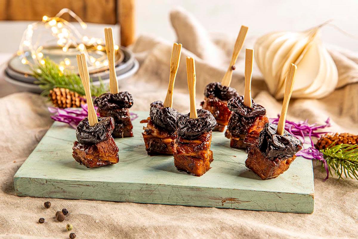 Biter av soyaglasert ribbe med sviske på toppen, holdt sammen med pinne. Bitene står på en lys grønn trefjøl, med granbar rundt. Julelys og julepynt i bakgrunnen.