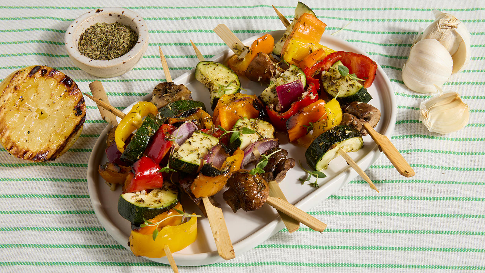 Grillspyd med grønnsaker servert på hvitt fat på grønnstripet duk. Grillet sitron og hel hvitløk ved siden av.