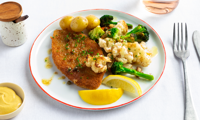 Ostepanetter av svin med grønnsaker og sitronsmør servert på en hvit tallerken med brokkolini, blomkål og små poteter. Tallerkenen står på hvitt middagsbord.
