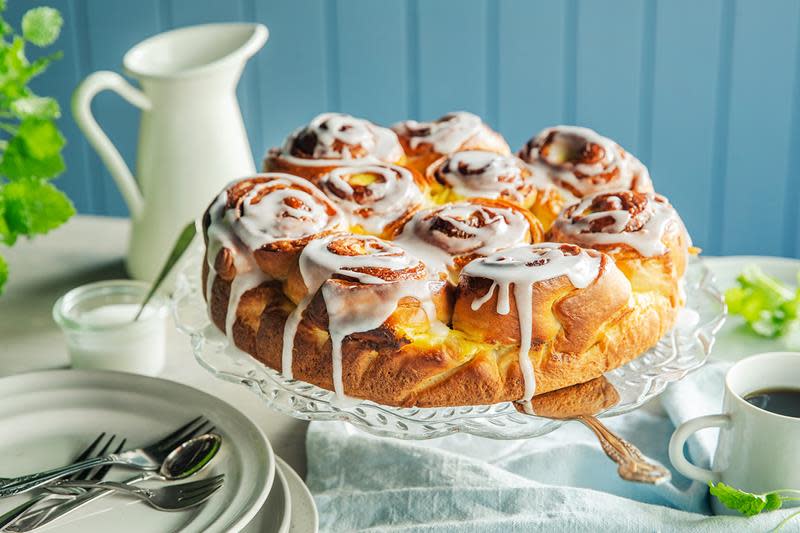 Gjærbaksten prinsessekake, med vaniljekrem i svingene, pyntet med melisglasur. Servert på stettfat, på hyggelig pyntet kaffebord. Blå bakgrunn.