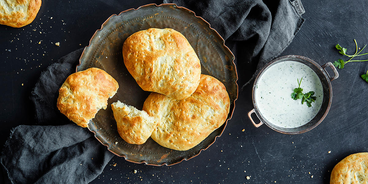 Saritas beste nanbrød servert på mørkt metallfat på mørkt bord med grå linserviett. Liten skål med raita ved siden av.