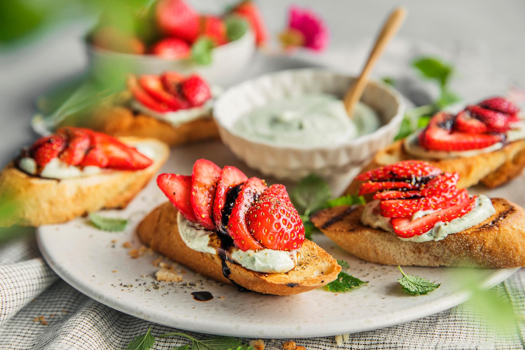 Bruschetta med jordbær og basilikumkrem servert på lys tallerken med en skål basilikumkrem i midten. Friske urter rundt, og en skål jordbær i bakgrunnen.