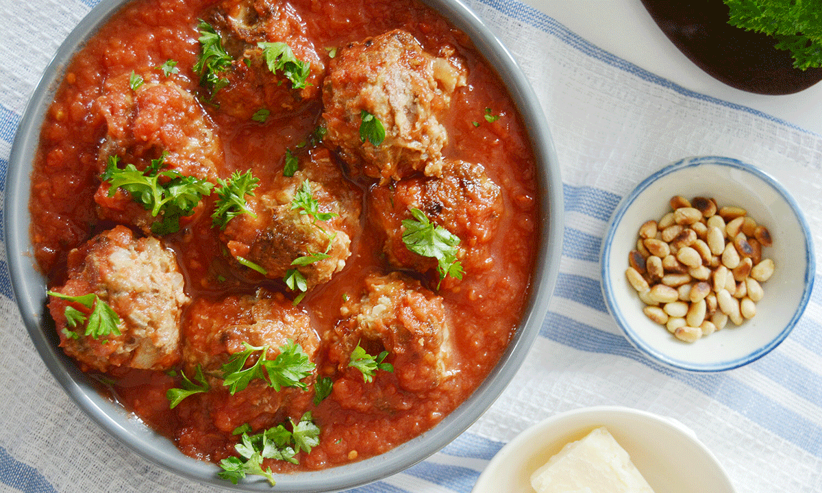 Spanske kjøttboller i tomatsaus, i skål pyntet med koriander ved siden av en skål med pinjekjerner på et hvitt og blått håndkle.