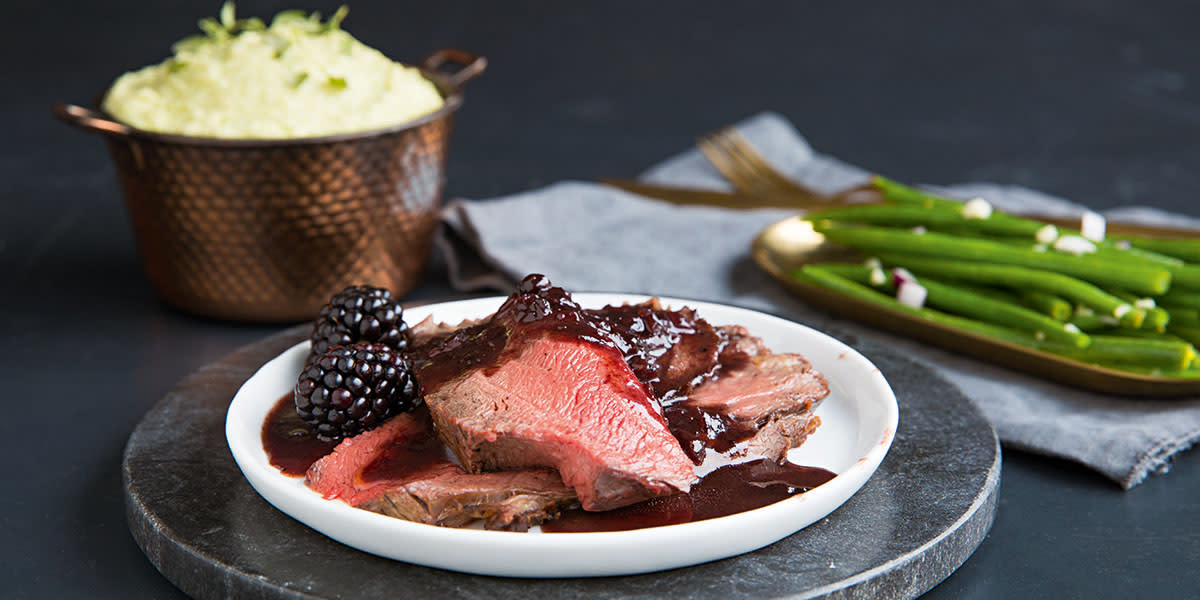 Saftig elgstek i skiver med solbærsaus og friske bjørnebær servert på hvit tallerken på mørkt steinfat. En messingskål med persillerotmos i bakgrunnen, og messingfat med grønne aspargesbønner på siden.