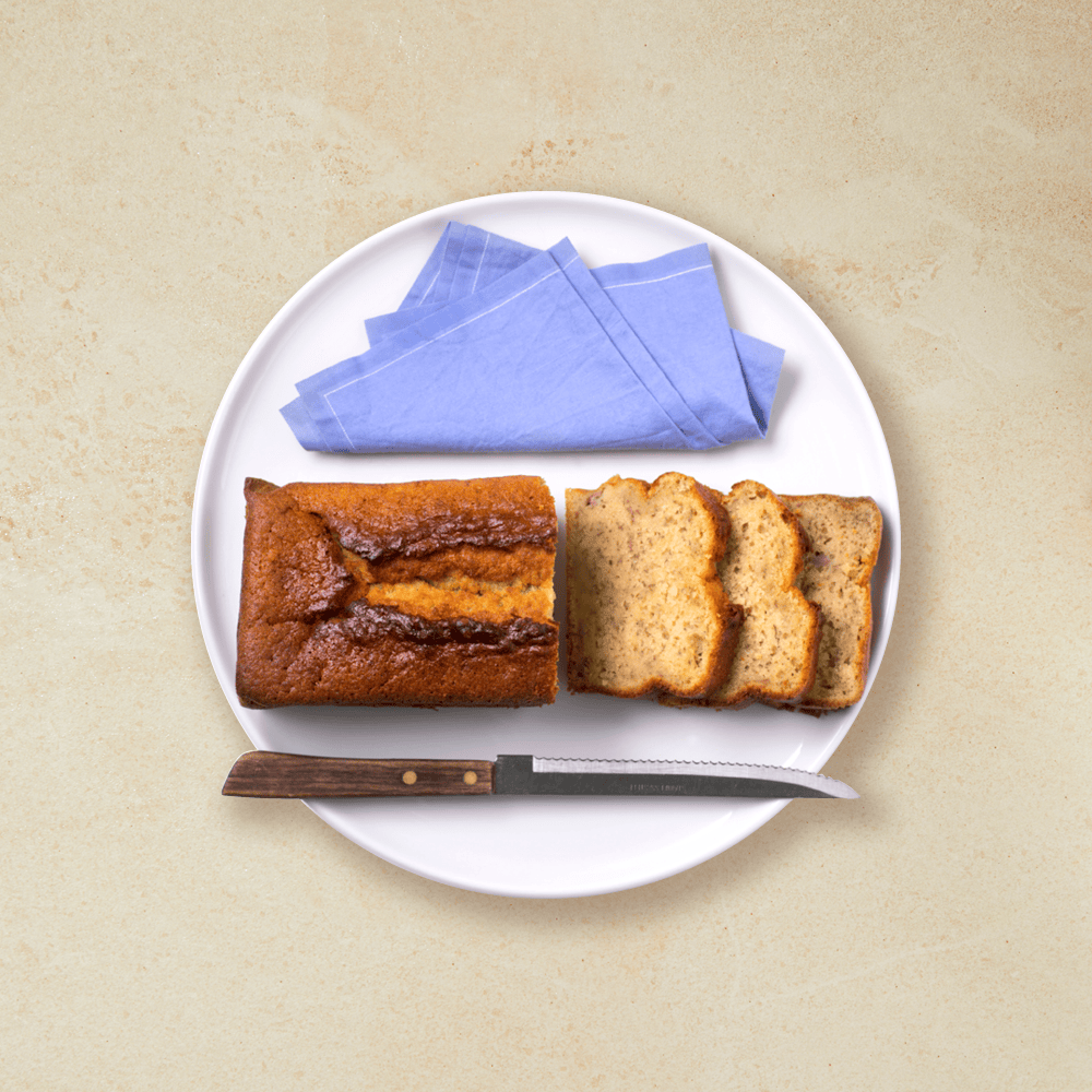 Enkel banankake skåret i skiver og servert på hvit asjett med lys lilla serviett ved siden av.