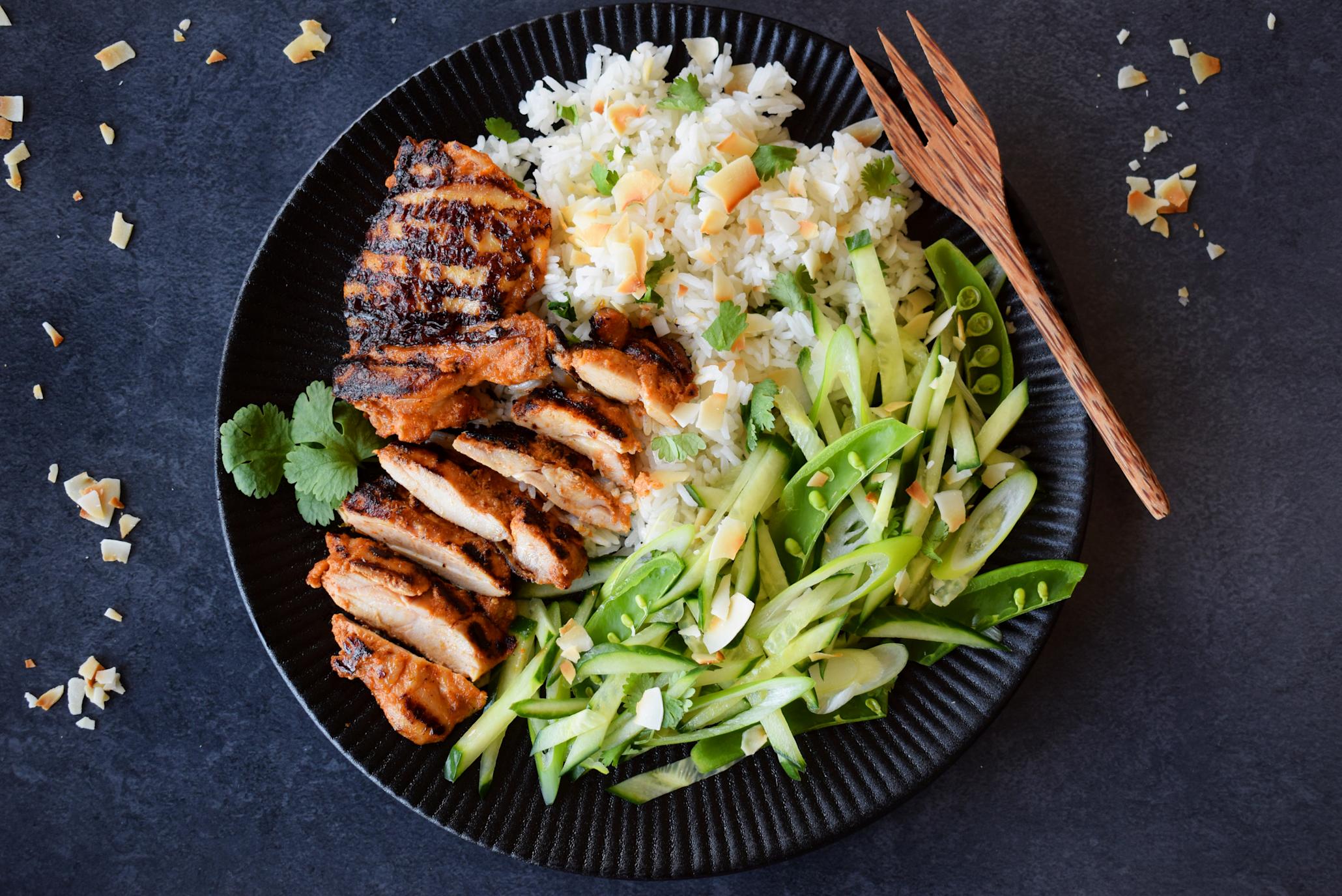 Grillet red curry kylling med koriander-ris, røstede kokosflak og grønn salat av agurk, sukkererter og vårløk, servert på svart tallerken og mørk bakgrunn. Gaffel i naturtre på siden.