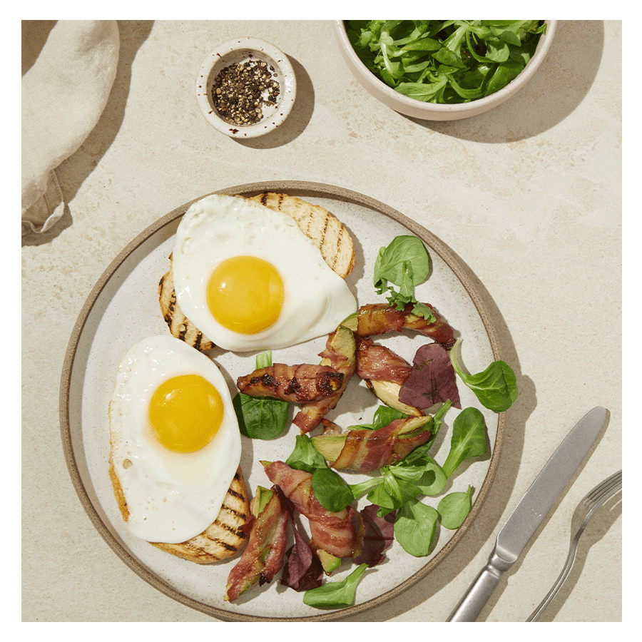 Speilegg på toast med baconsurret avokado i skiver, servert på lys tallerken på beige bakgrunn med bestikk ved siden av. Liten skål med salatmiks på siden.