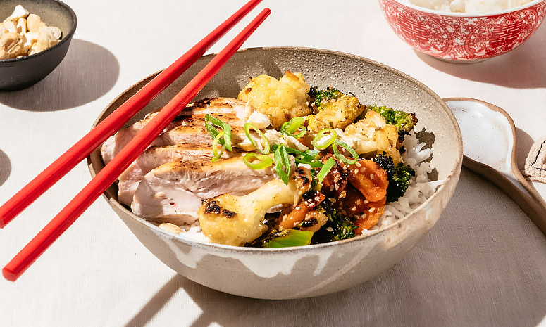 Kylling med ris brokkoli og sesamfrø servert i en dyp beige skål med røde spisepinner på kanten, ligger på en beige dyk