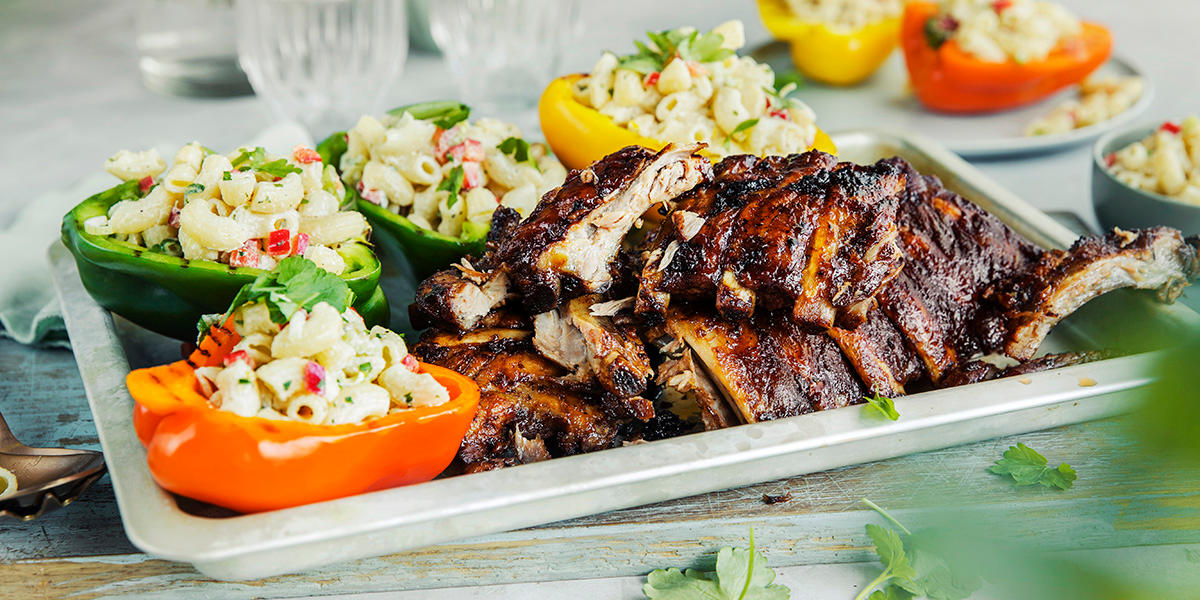 Saftige spareribs med fargerik makaronisalat i halve paprikaer servert på stekebrett med friske urter på toppen. Brettet står på dekket middagsbord i tre.