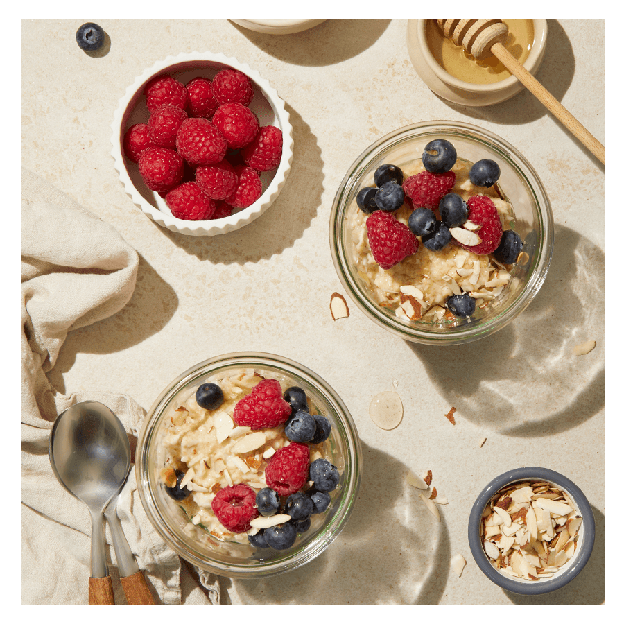 Kjøleskapsgrøt med havregryn servert i glass med friske bringebær, blåbær, mandler og honning. Små skåler med friske bringebær, skivede mandler og honning rundt.