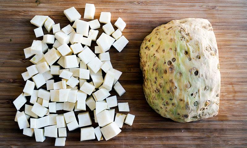 Sellerirot delt i terninger ved siden av en halv sellerirot, på mørkt trebord.