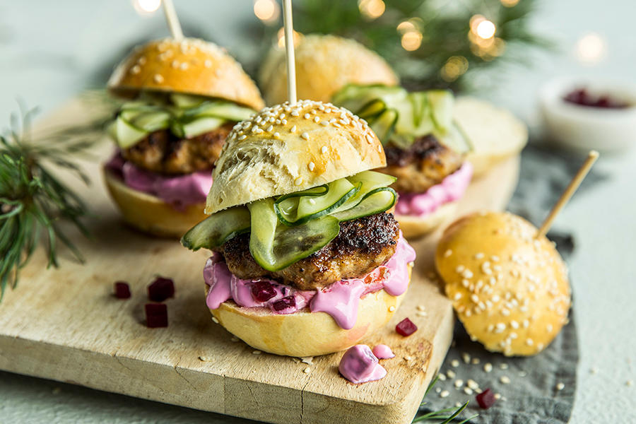 Sliders med medisterkaker, syltet agurk og rødbetekrem servert på lys trefjøl med granbar og julelys i bakgrunnen. Hver minihamburger har en tannpirker stukket gjennom.