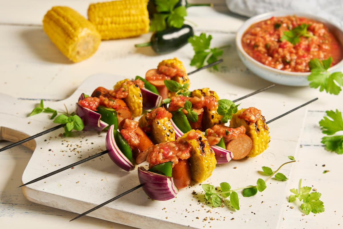 Grillspyd med chicken sausage og salsa roja, skiver av maiskolber, rødløk og grønn paprika servert på hvit trefjøl. Maiskolber, jalapeno, koriander og en skål salsa roja i bakgrunnen.