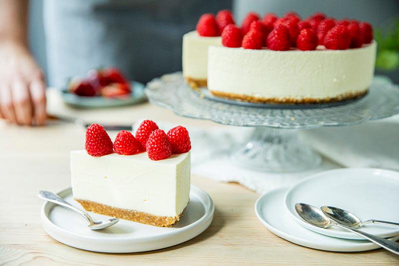 Klassisk ostekake pyntet med bringebær og servert på kakefat i glass på dekket bord med asjetter og teskjeer.