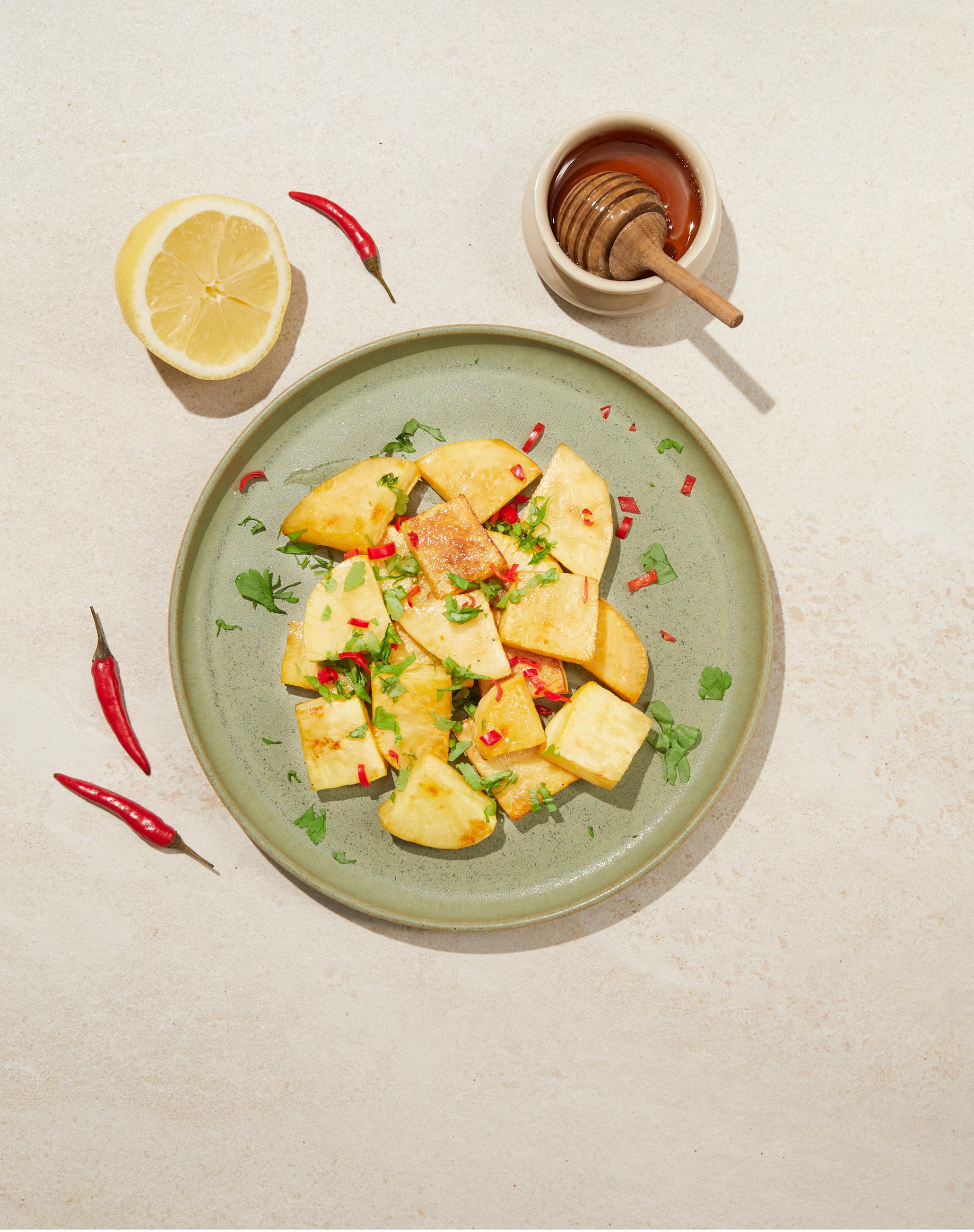 Kålrot med honning og chili, servert i små skiver på lys grønn tallerken på beige bakgrunn. Liten skål med honning ved siden av, samt en halv sitron og små ferske chili. Ferske urter strødd over tallerkenen.