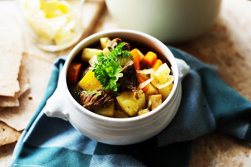 Brun lapskaus med fargerike grønnsaker, servert i liten hvit suppebolle, med flatbrød og smør på siden. Blå linserviett og rustikk trebakgrunn.