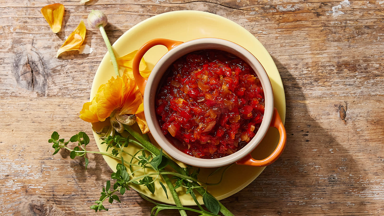 Paprikarelish i liten oransje skål med blomster og friske urter rundt.