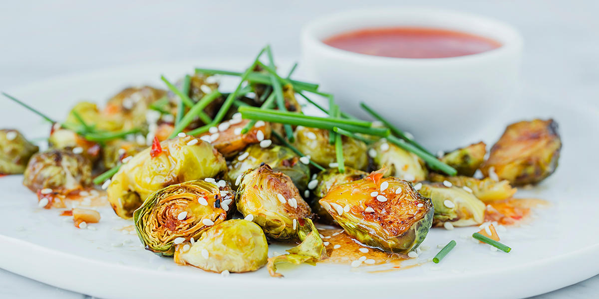 Glasert rosenkål med sweet chili, delt i to og servert på hvit tallerken med sesamfrø, gressløk og en liten skål sweet chili saus.