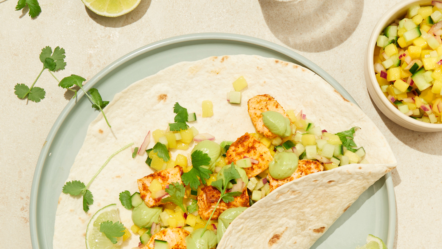 Fisketaco med hjemmelaget guacamole og mangosalsa servert i tacolefse på lyseblå tallerken, med frisk koriander og limebåter til. Liten skål med mangosalsa, og vannglass ved siden av.