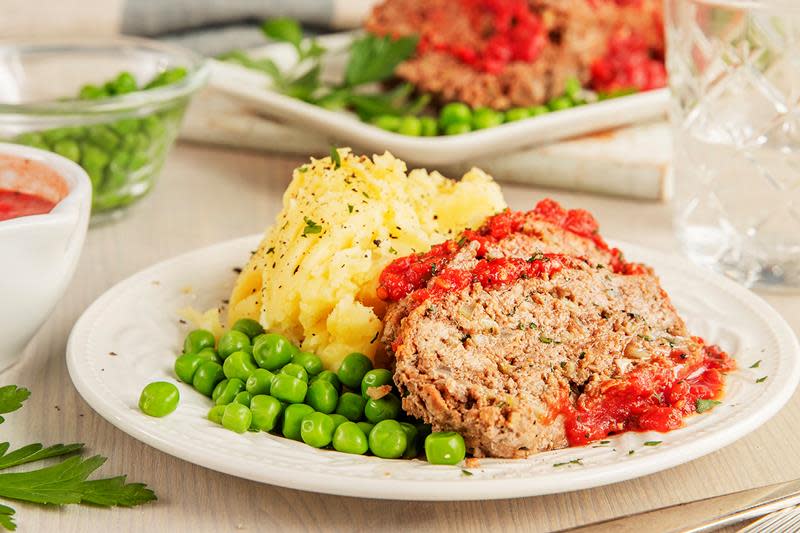 Hjemmelaget kjøttpudding med tomatsaus på toppen, servert på hvit tallerken med potetmos og grønne erter. Lyst middagsbord.