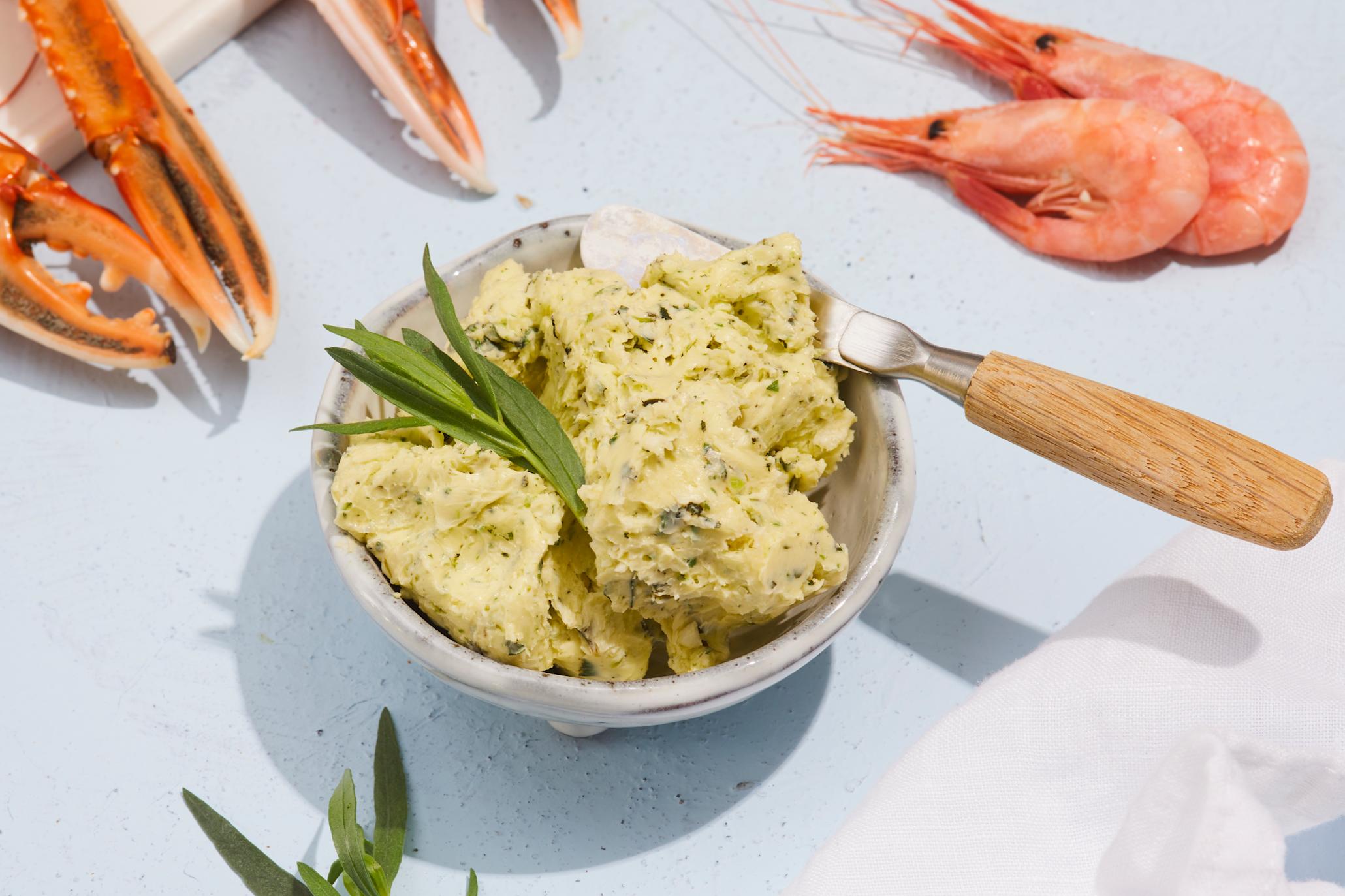 Hjemmelaget hvitløkssmør til skalldyr servert i liten keramikkskål på lyseblått bord med friske reker og sjøkreps. Frisk estragon på toppen.
