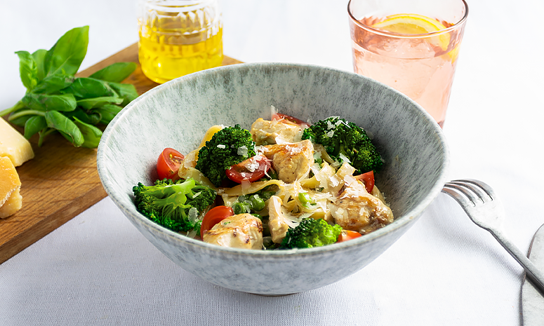 kylling med kremet tagliatelle og grønnsaker i en grå skål med en skjærehøvel, og to glass i bakgrunnen og kniv og gaffel ved siden av