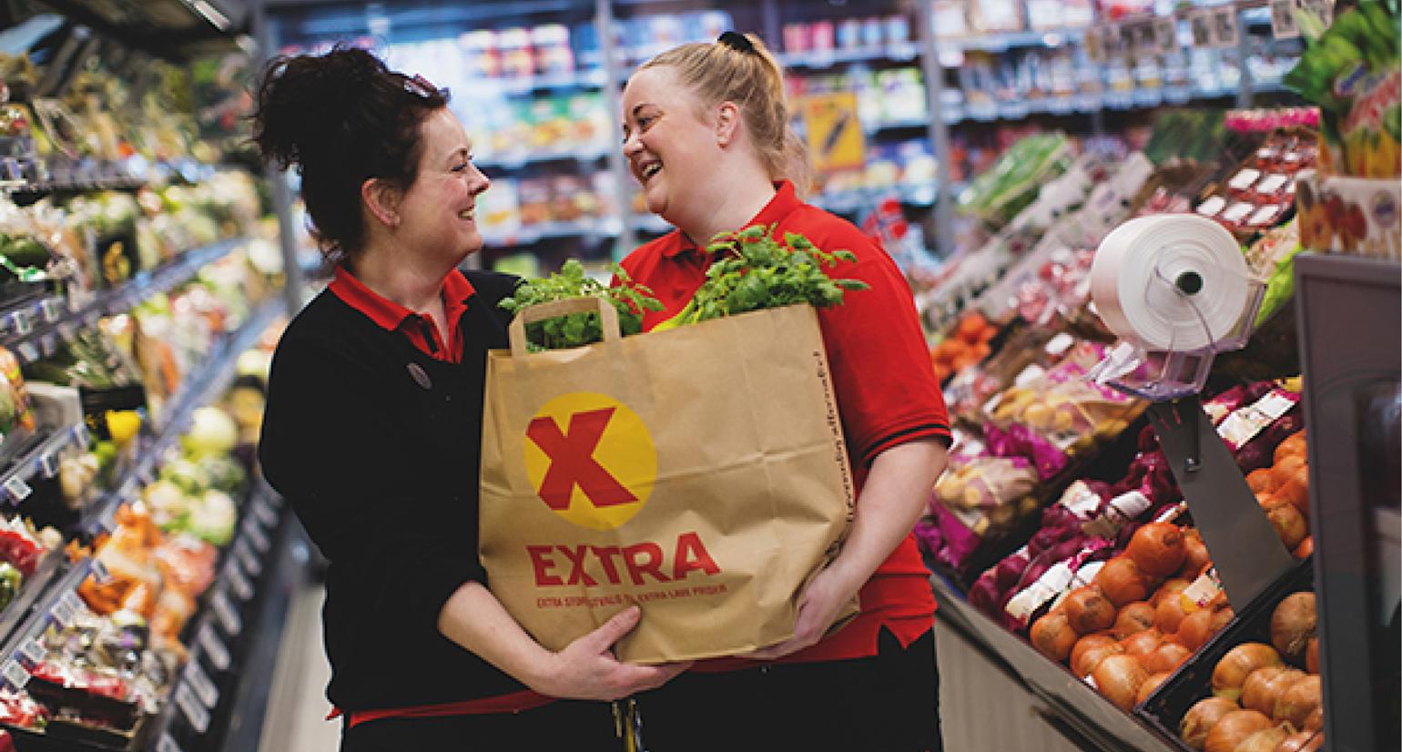 To damer holder i en papir Extra poser med grønnsaker og smiler.