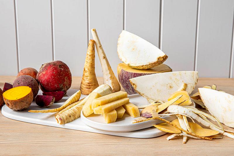 Fargerike rotgrønnsaker av forskjellige slag, oppdelt og hele, på hvit marmorfjøl på et bord. Skrell og biter av rotgrønnsaker ved siden av.