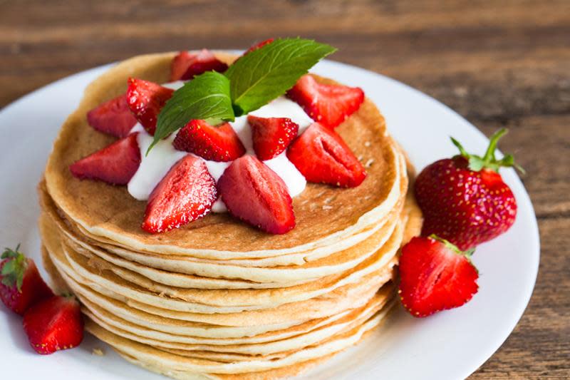 Stabel med nystekte pannekaker på hvit tallerken, toppet med yoghurt og ferske jordbær. Rustikk trebakgrunn.